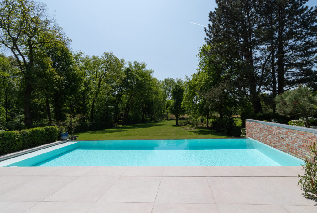 Panorama-Ansicht eines langen Infinity-Pools mit hellblauer Auskleidung, einer flachen Einstiegszone und hellen Terrassenplatten in einem weitläufigen Garten